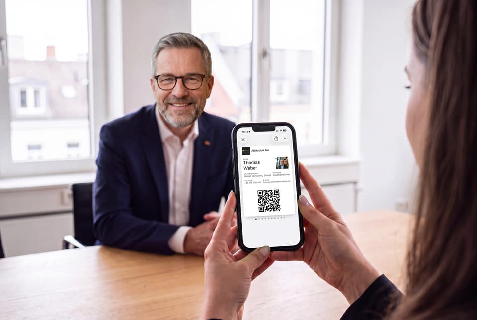 Mit KI gestaltete Szene: Thomas Weber, fiktive Person, sitzt bei Tageslicht am Besprechungstisch; im Vordergrund hält eine Kundin ihr iPhone mit seiner Karte im Wallet.